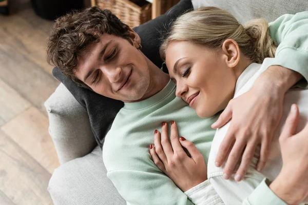 Top view of happy young man embracing girlfriend while lying on couch in living room — Fotografia de Stock