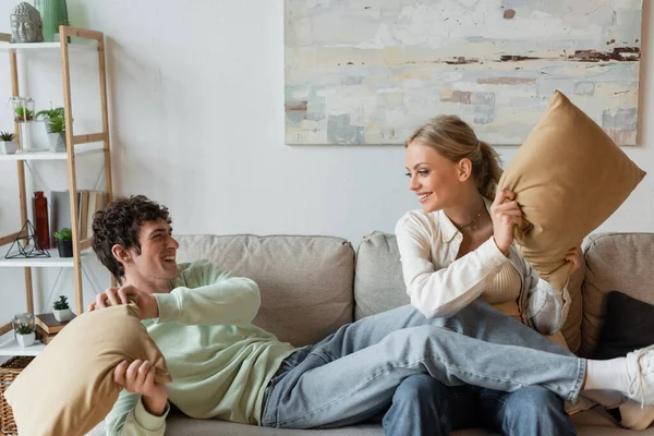 Positive young couple smiling while having pillow fight in living room — Stockfoto