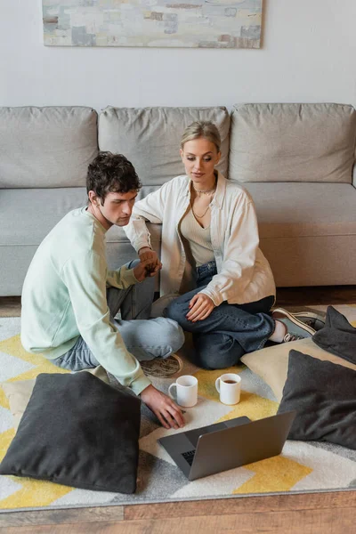 Curly man holding hand of blonde woman while looking at laptop near cups on carpet — стоковое фото