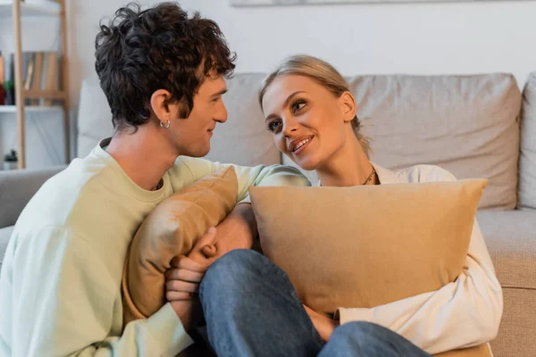 Cheerful young woman with blonde hair and curly man looking at each other while holding pillows - foto de stock