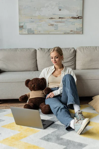 Молода жінка в джинсах тримає плюшевого ведмедя і дивиться фільм на ноутбуці — стокове фото