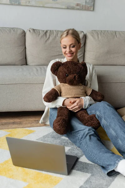 Positive young woman in jeans holding teddy bear and watching movie on laptop — стоковое фото