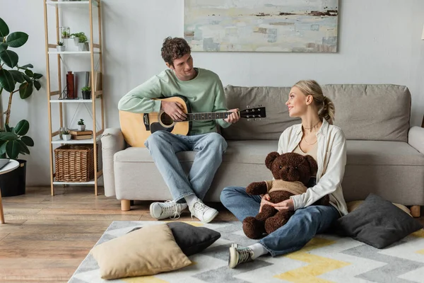 Curly man sitting on couch and playing acoustic guitar near happy woman with teddy bear — Photo de stock