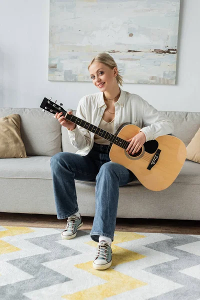 Positive and blonde woman playing acoustic guitar while sitting on couch — Photo de stock