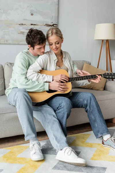 Happy blonde woman playing acoustic guitar with curly boyfriend while sitting on couch — Stock Photo