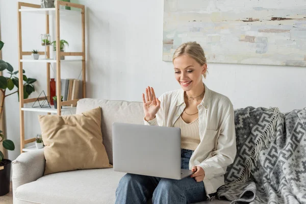 Cheerful young woman with blonde hair waving hand during video call on laptop — Foto stock