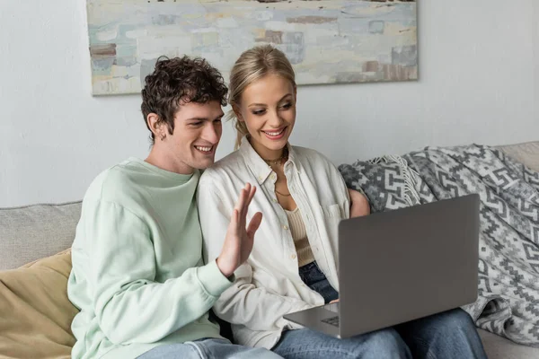 Cheerful young man with curly hair waving hand near girlfriend during video call on laptop — Stock Photo