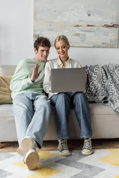 Happy young man with curly hair waving hand near girlfriend during video call on laptop — Foto stock