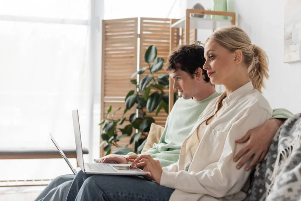 Side view of curly man and blonde woman using laptops while working from home in living room - foto de stock