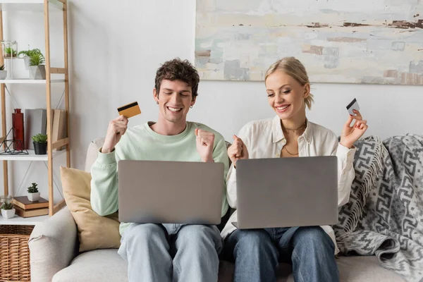 Happy young couple holding credit cards near laptops while doing online shopping — Stock Photo