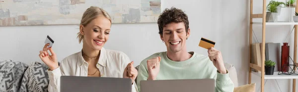 Happy young couple holding credit cards near laptops while doing online shopping, banner — Stock Photo