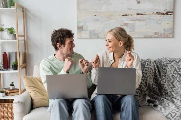 Excited young bloggers rejoicing near laptops while sitting on couch - foto de stock