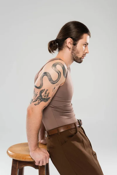 Side view of long haired and tattooed man posing near chair isolated on grey — Stock Photo