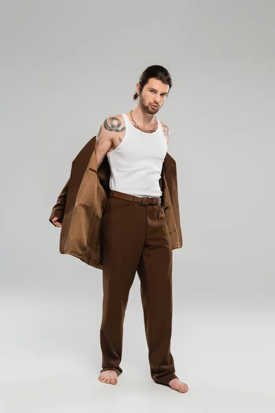 Full length of barefoot tattooed man taking off jacket on grey background — Stock Photo
