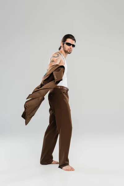 Full length of stylish tattooed man in sunglasses holding jacket on grey background - foto de stock