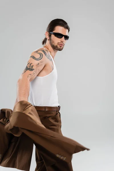 Trendy tattooed man in sunglasses holding jacket isolated on grey — Stockfoto