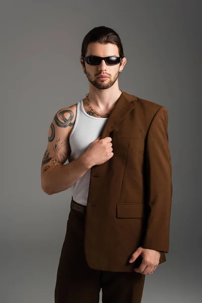 Bearded tattooed man in sunglasses holding jacket isolated on grey — Stock Photo