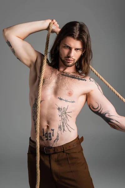 Shirtless tattooed model posing with rope and looking at camera isolated on grey — Stock Photo
