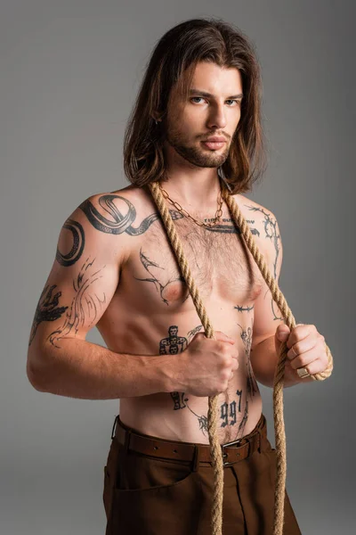 Сексуальна татуйована модель позує з мотузкою і дивиться на камеру ізольовано на сірому — стокове фото