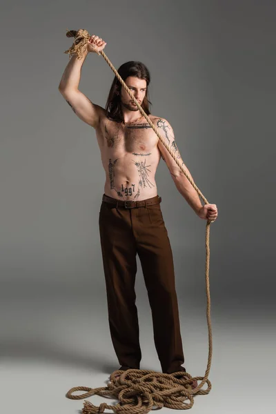 Full length of tattooed model in pants holding rope on grey background - foto de stock