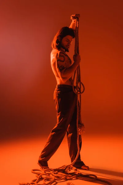 Повна довжина сексуальної татуйованої моделі позує з мотузкою на червоному тлі — стокове фото