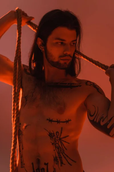Young tattooed and shirtless model posing with rope isolated on red — Stock Photo
