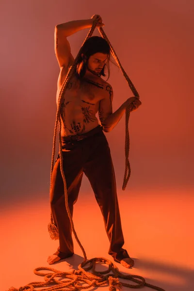 Full length of sexy tattooed model posing with rope on red background - foto de stock