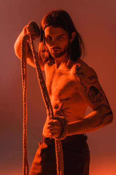 Long haired and tattooed model holding rope and looking at camera on red background - foto de stock