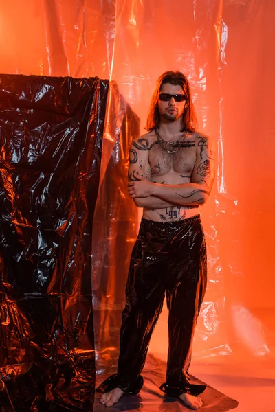 Tattooed man in latex pants and sunglasses standing near cellophane — стоковое фото