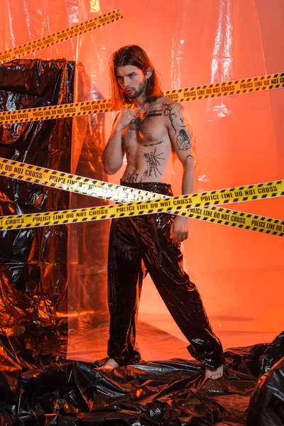 Tattooed man in latex pants standing near police line and cellophane - foto de stock