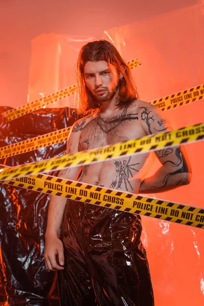 Long haired and tattooed man looking at camera near police line and polyethylene at background — Stock Photo
