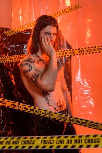Shirtless tattooed model touching face near police line and polyethylene at background — Stock Photo