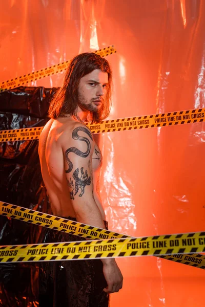 Shirtless tattooed model looking at camera near police line and cellophane at background — Stock Photo