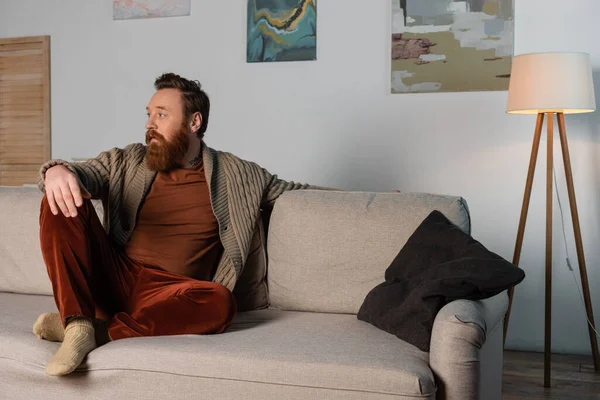 Bearded man in cardigan sitting on couch in living room — Photo de stock