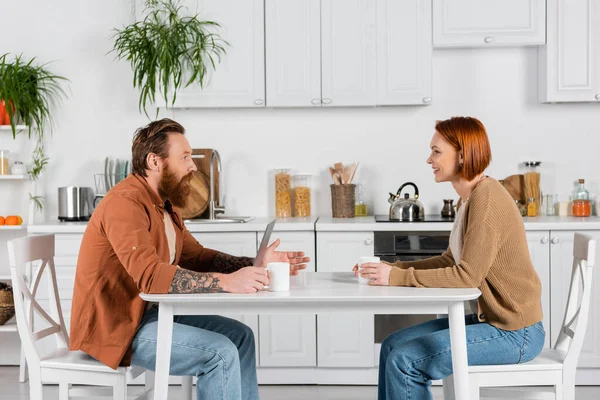 Side view of bearded man talking to redhead wife near laptop and cups in kitchen — Stock Photo