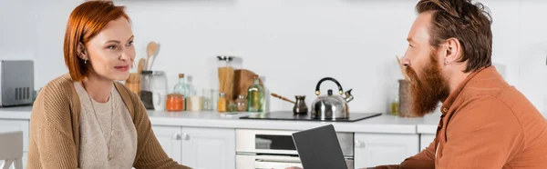Smiling redhead woman looking at bearded husband near laptop in kitchen, banner — Stock Photo