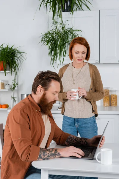 Redhead woman holding cup while husband using laptop in kitchen — Stock Photo