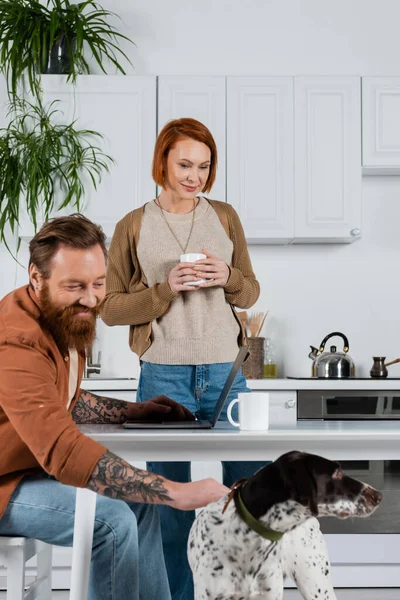 Cheerful tattooed man petting dalmatian dog near laptop and wife with cup in kitchen — Photo de stock