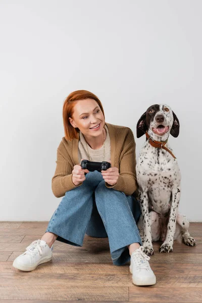 KYIV, UKRAINE - DECEMBER 16, 2022: Cheerful red haired woman holding joystick and looking at dalmatian dog at home — Photo de stock