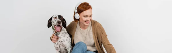 Femme souriante dans des écouteurs sans fil étreignant chien dalmate à la maison, bannière — Photo de stock