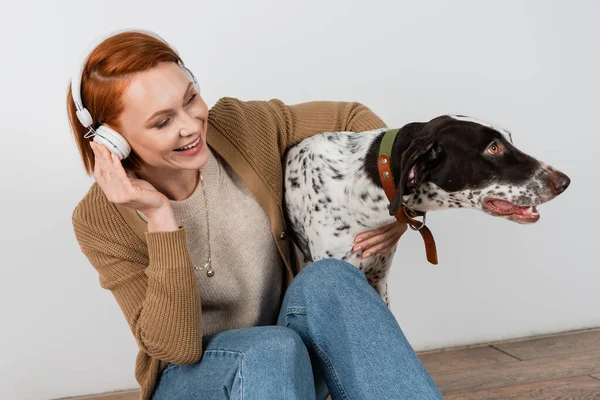 Cheerful redhead woman listening music in headphones and hugging dalmatian dog at home — Stock Photo