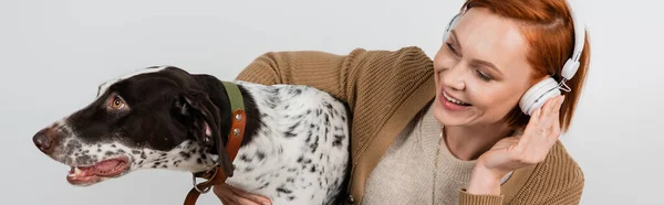 Femme positive dans les écouteurs étreignant chien dalmate isolé sur gris, bannière — Photo de stock