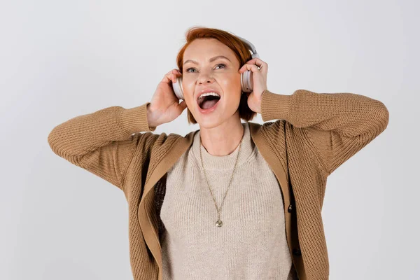 Excited and red haired woman in headphones singing isolated on grey — Foto stock
