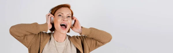 Excited and red haired woman in headphones singing isolated on grey, banner — Foto stock