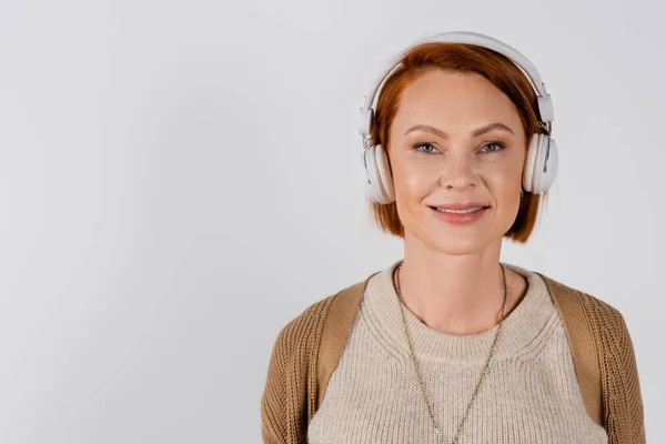 Femme rousse souriante écoutant de la musique dans des écouteurs isolés sur gris — Photo de stock