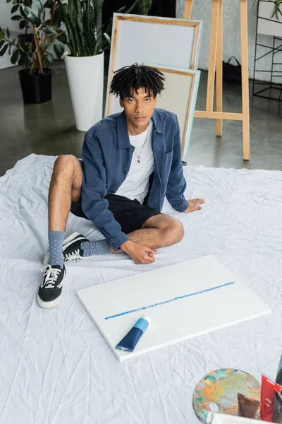 Young african american artist looking at camera while sitting near paints and canvas in workshop — Stock Photo