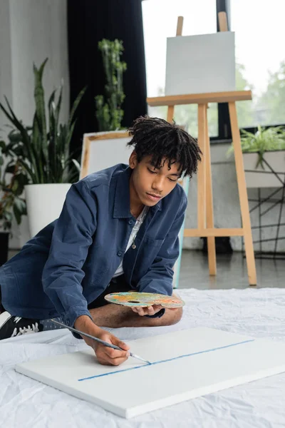 Young african american artist painting on canvas while sitting on cloth in studio — Stock Photo