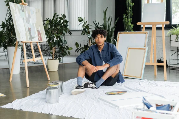 African american artist looking at camera while sitting near paints and canvas on floor in workshop — Fotografia de Stock