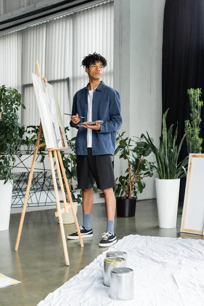 Young african american artist holding paintbrush and palette near canvas on easel in studio — Stock Photo