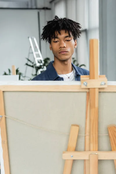 Young african american artist looking at blurred canvas on easel in workshop — Fotografia de Stock